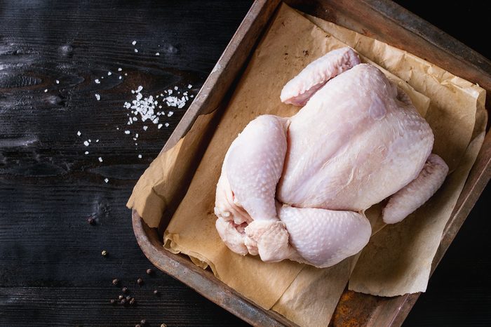 Raw organic uncooked whole chicken with salt and pepper on backing paper in old oven tray over black burnt wooden background. Top view with space.