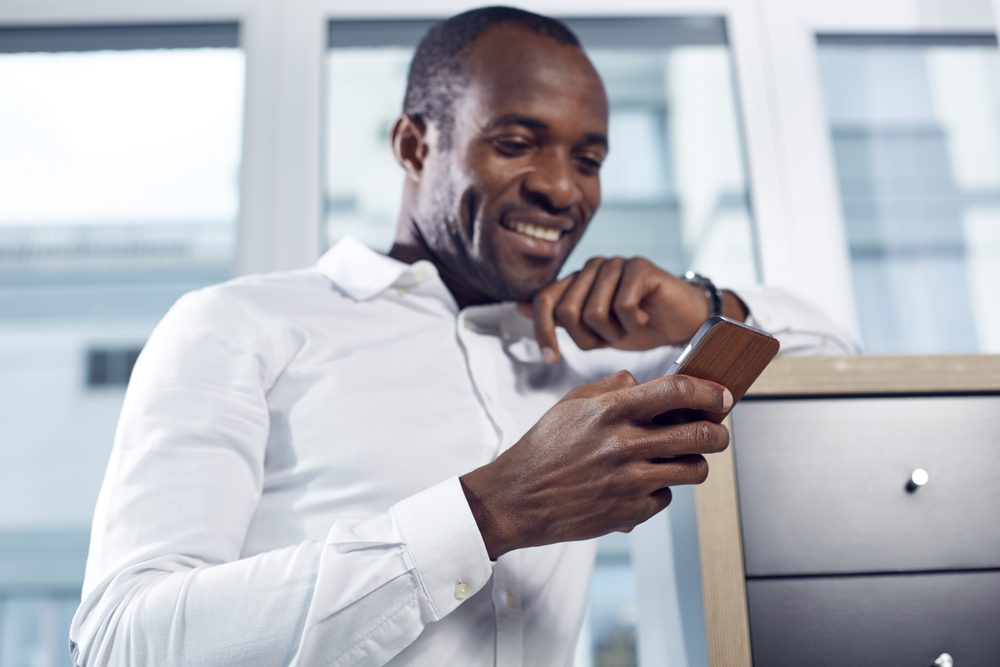 Optimistic news. Selective focus of hand with mobile phone of positive young professional businessman. He is standing in office while leaning elbow on shelf and looking at screen of gadget with smile