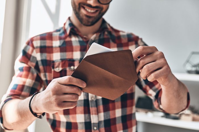 Receiving greeting card. Close up of young man opening envelope and smiling while standing indoors