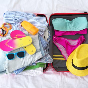 Open suitcase with different clothes and accessories for summer journey on bed