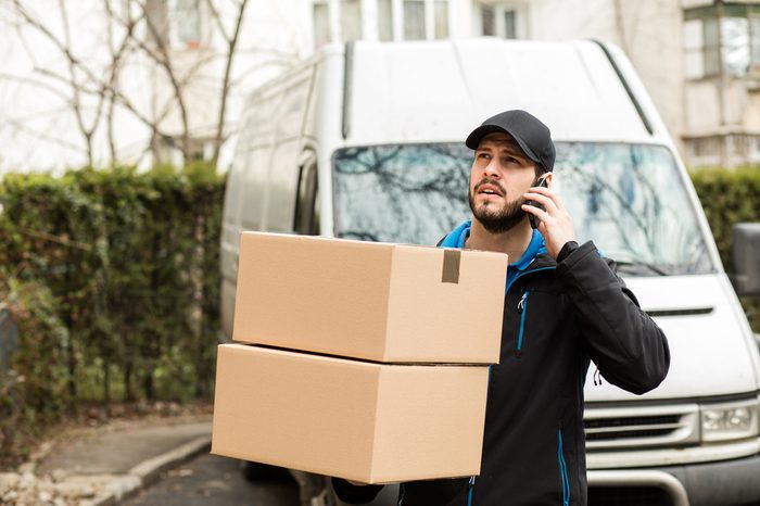 Delivery man with cap and cardboard in hands, talking at cellphone having his van behind