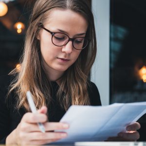 Portrait of a young beautiful girl sitting at table with laptop, holding pen in hand  and reading piece of paper. Education concept