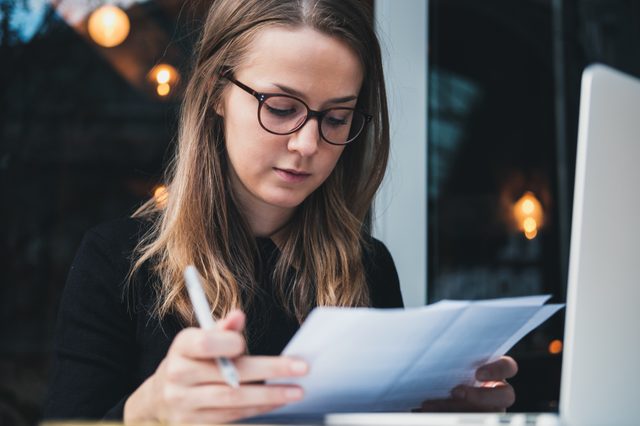 Portrait of a young beautiful girl sitting at table with laptop, holding pen in hand  and reading piece of paper. Education concept
