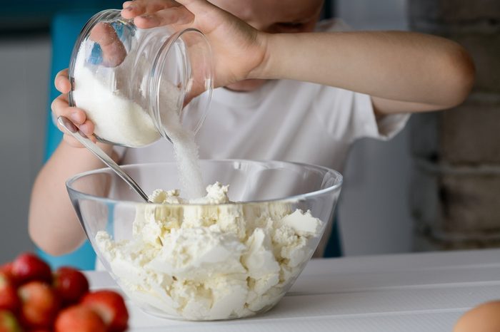 7 year old boy adding sugar to cottage cheese in a bowl. Prepares mini cheesecakes with strawberries