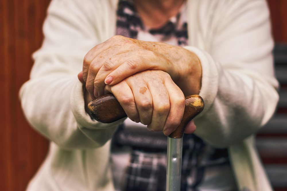 Senior woman sitting outside on a wooden bench and rests. Hands of a woman pensioner holding a walking stick.