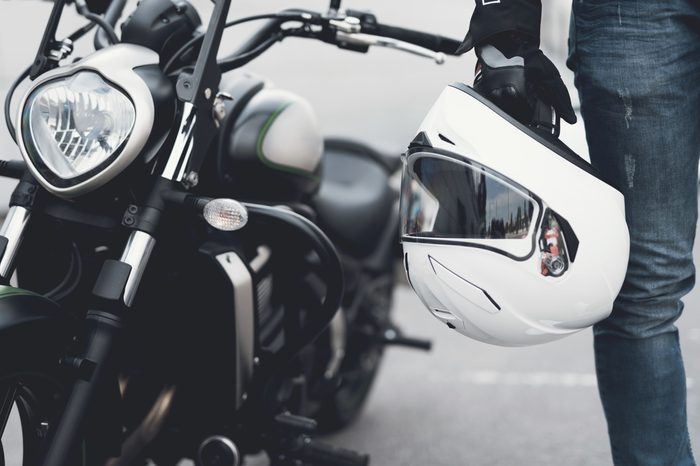 A young guy stands near an electric motorcycle. He is wearing jeans and is holding a helmet.