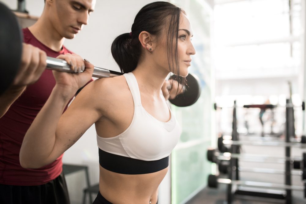 Waist up portrait of fitness coach helping beautiful young woman training with barbell in modern gym, copy space