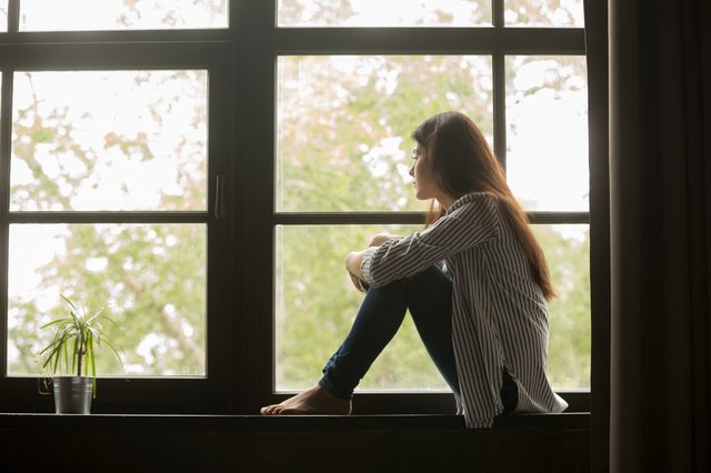 Thoughtful girl sitting on sill embracing knees looking at window, sad depressed teenager spending time alone at home, young upset pensive woman feeling lonely or frustrated thinking about problems