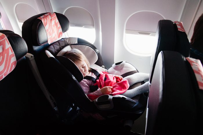 Flying with children: two year old baby sleeping in her own car seat setting on an ordinary seat on a commercial airliner. Concept photo of air travel with baby. Natural in-plane lightning conditions