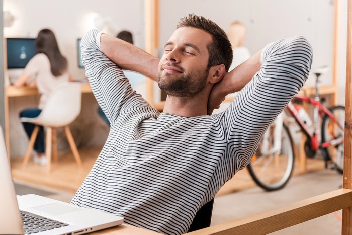 Taking time for a minute break. Cheerful young man holding hands behind head and keeping eyes closed while sitting at his working place with his colleagues working in the background