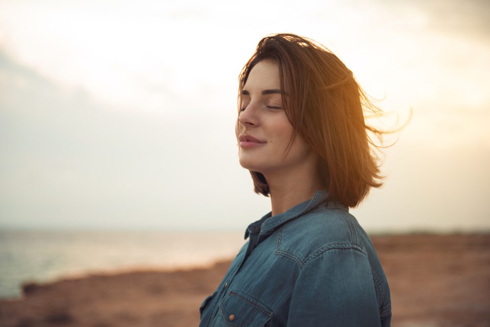 Great pleasure. Charming calm young woman is standing near sea with closed eyes and expressing delight. She is posing against wonderful sunset while enjoying last rays of the sun