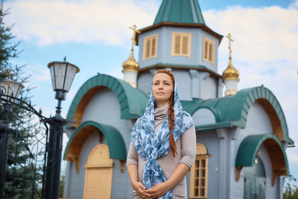 Russian Orthodox middle-aged woman in a scarf stands on the background of the Orthodox wooden Church.