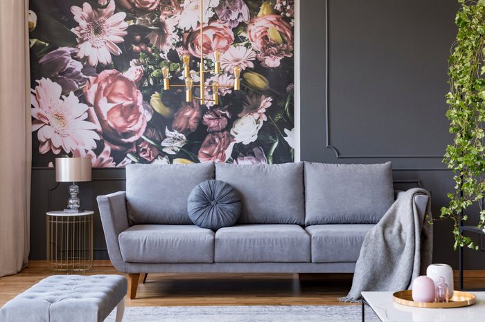 Blanket on grey couch in living room interior with flowers wallpaper and lamp on table. Real photo