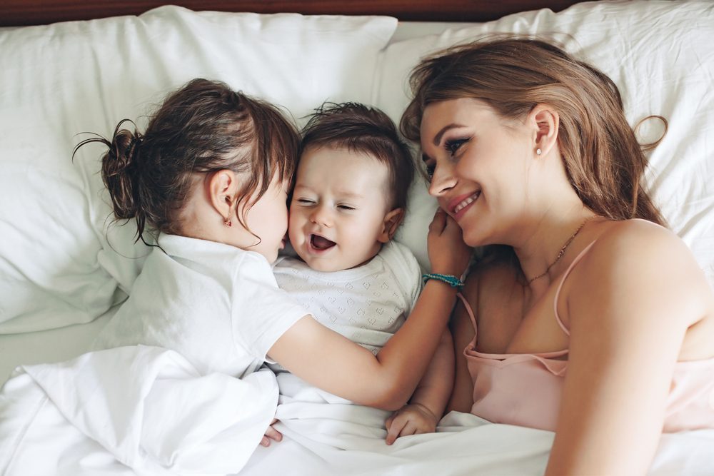 Young mom with her 5 years old daughter and 4 months old baby dressed in pajamas are relaxing and playing in the bed at the weekend together, lazy morning