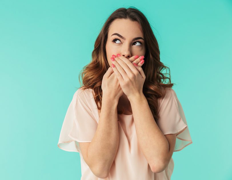 Portrait of a lovely young girl in dress standing with mouth covered and looking away at copy space isolated over blue background