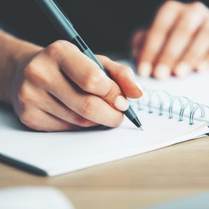 Close up of woman's hands writing in spiral notepad placed on wooden desktop with various items