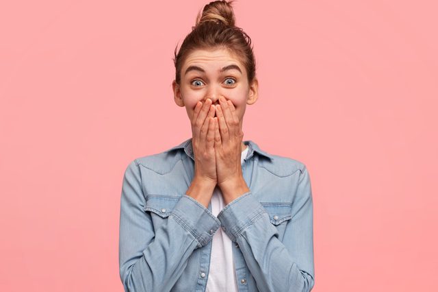 Young vivacious beautiful young female giggles joyfully, covers mouth, has fun alone, stands against pink background or studio wall, has natural laughter, hears positive story or funny anecdote