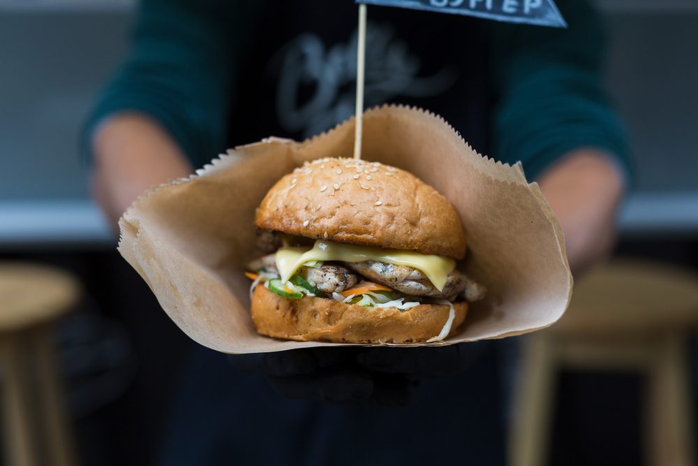 Fresh burger cooked at barbecue outdoors in craft paper. Big hamburger with steak meat and vegetables closeup, chef unfocused at background. Street fast food.