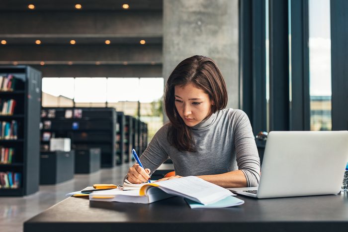 Female student taking notes from a book at library. Young asian woman sitting at table doing assignments in college library.