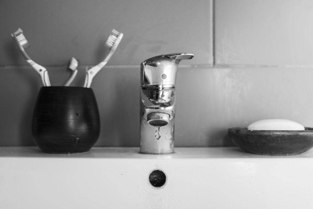 Scarce water/Black and white image of a bathroom sink with the tap dripping a drop of water, two normal toothbrushes plus a small one and a piece of soap in a soap dish.