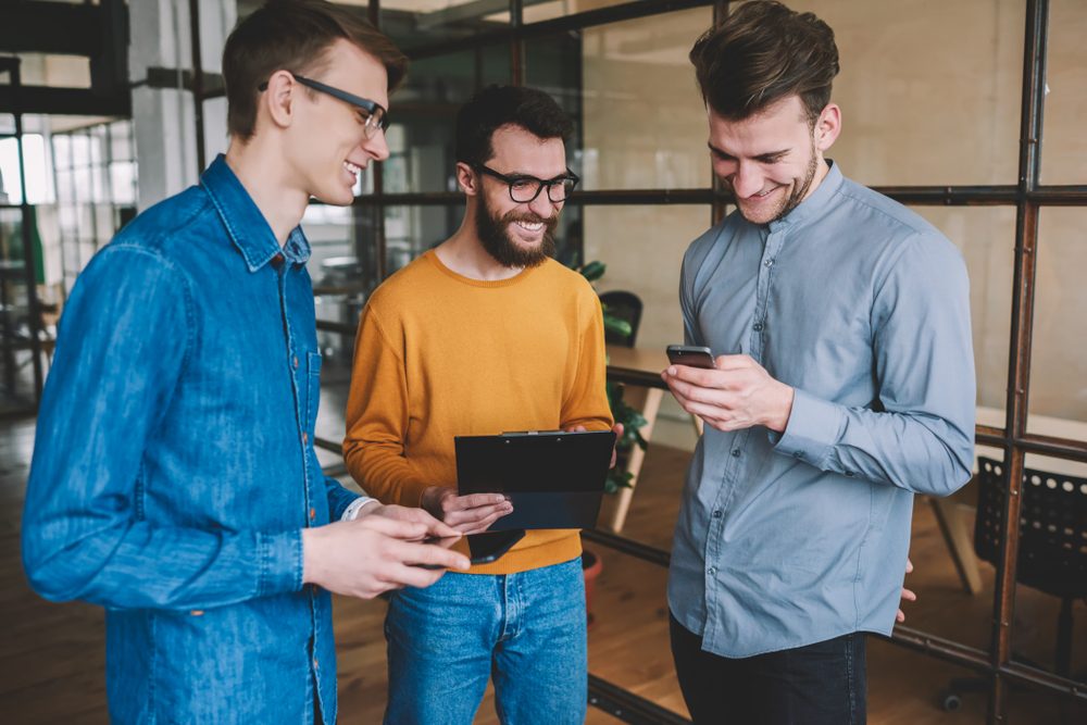 Successful male partners enjoying break of work and laughing from funny joke reading in social network via smartphone device, young positive men communicated and using technology in modern office