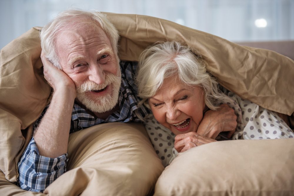 Cheerful old married couple lying in bed under blanket. Woman is laughing and man is looking at camera with smile. Concept of happiness
