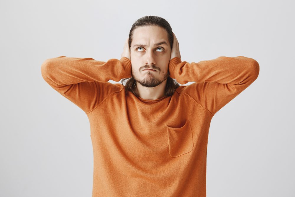 Neighbors arranged loud party. Portrait of angry annoyed young man with beard, holding up, covering ears with palms, being in rage from hearing disturbing noise from upstairs, standing over gray wall