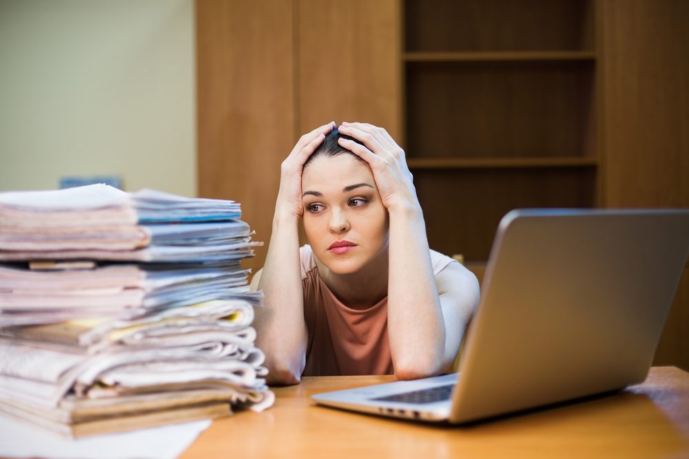 A young woman sits in front of a pile of papers and a computer holding her head