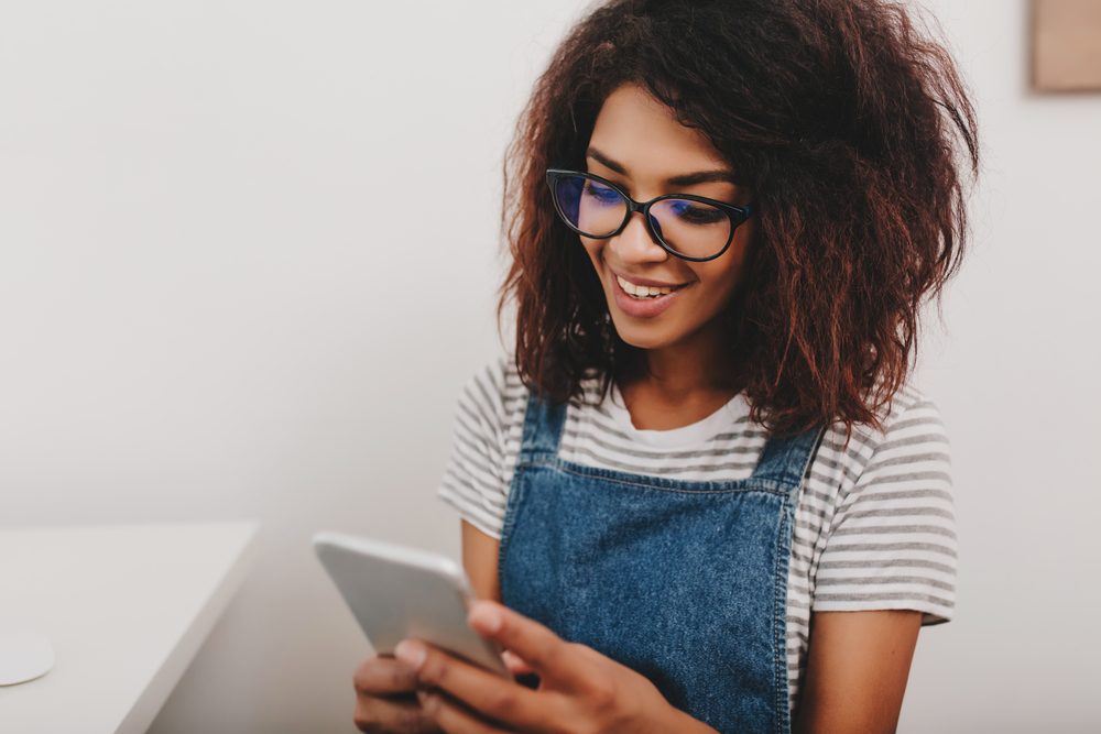 Cute lady with bronze skin checking new messages in social networks using her phone. Indoor portrait of smiling curly girl testing mobile app on smartphone sitting on light background.