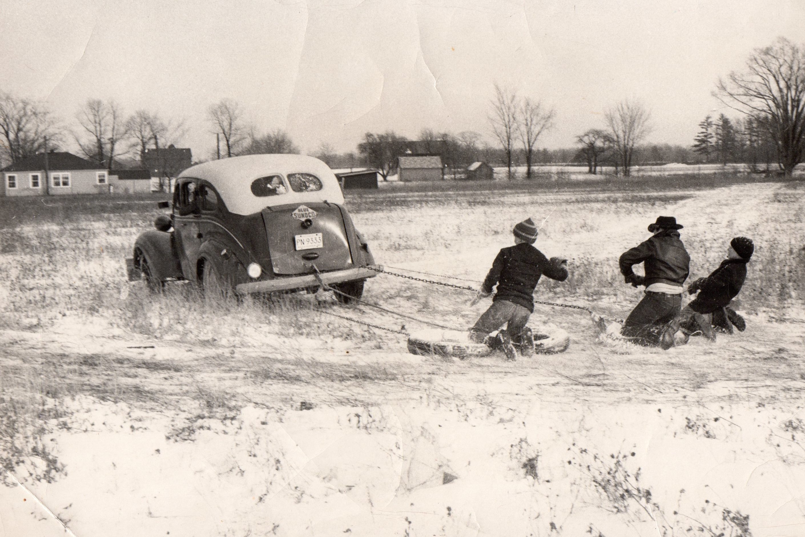 This scene takes place in Goodells; MI. about 1958. The car belonged to my father; Sandy Sanderson. He used the 1937 Dodge with a floor shift as a service car for his gas station (note the large front wooden bumper) to push-start cars. He was inspired to let any of the neighbor kids learn to drive in the field by using his service auto. Most of us learned to drive by 12 to 16 yrs. The object in the winter-the driver was to try to dump anyone riding. When you got cold; places were changed with someone inside the car. The person in the middle was Don Schattler (now deceased) the other two are the Polivich brothers. We drove the car in the field next to my fathers gas station; the tires we rode on; ropes; and chains were furnished by him.