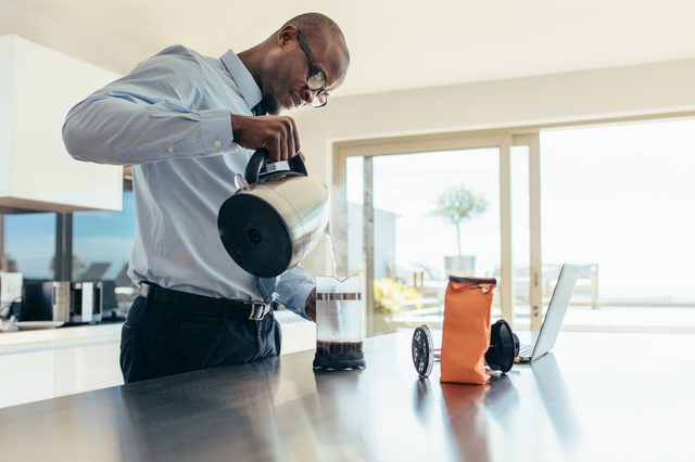 Man pouring hot water in coffee maker. Businessman preparing coffee on breakfast table with a laptop computer by the side.