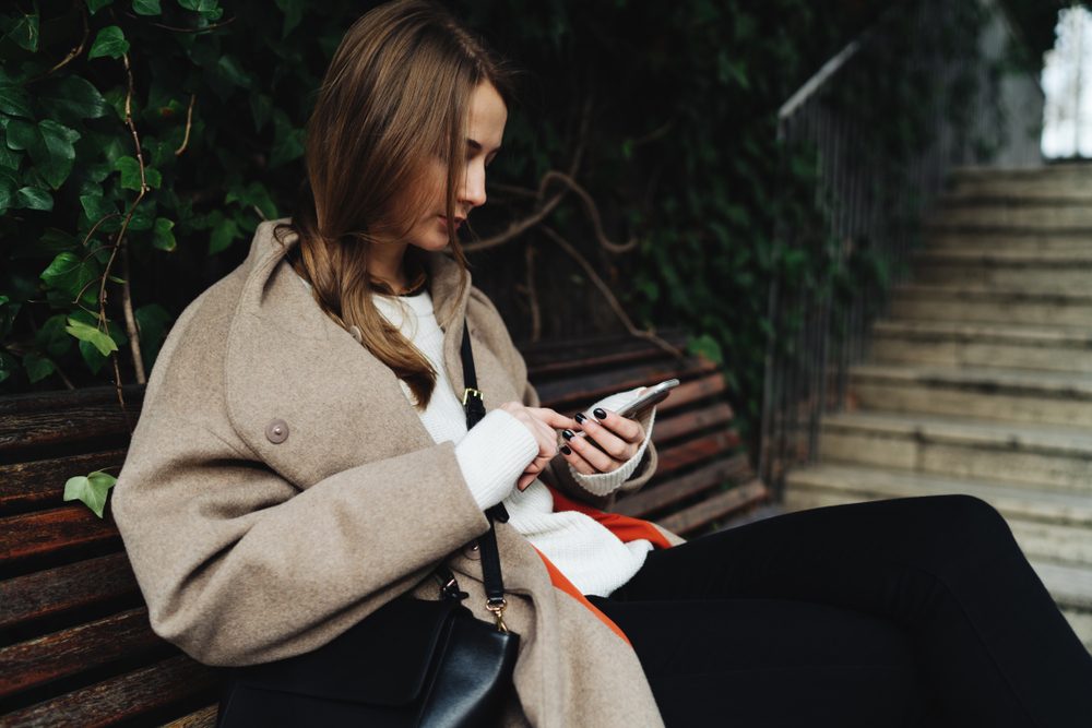 Portrait of blonde caucasian woman wearing trendy clothes holding mobile phone while sitting on a natural background. Student girl is surfing the web on a smartphone while relaxing in park.
