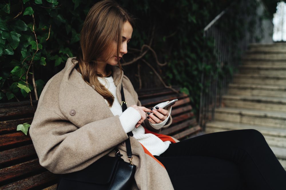 Portrait of blonde caucasian woman wearing trendy clothes holding mobile phone while sitting on a natural background. Student girl is surfing the web on a smartphone while relaxing in park.