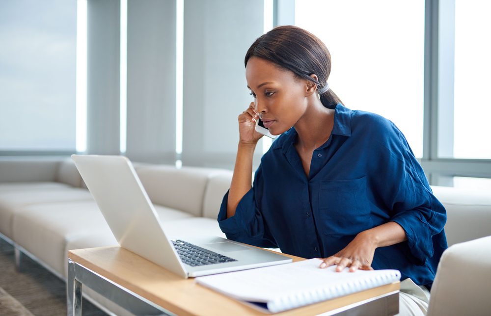 Focused young businesswoman talking on a cellphone and working on a laptop while sitting at a table in a modern office