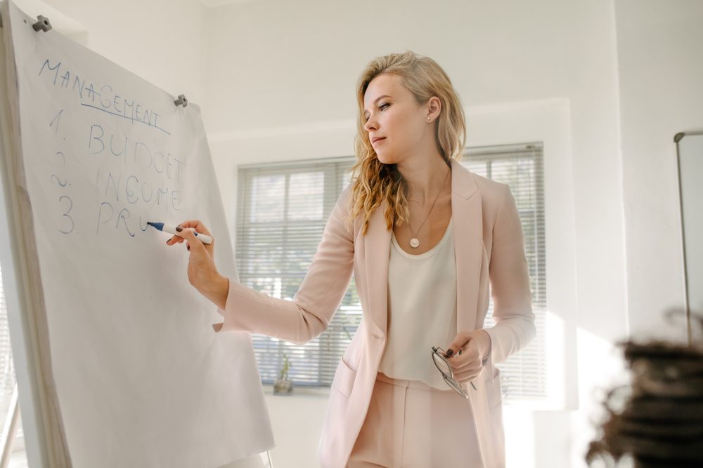 Female manager makes a presentation to colleagues in boardroom. Woman writing on flip chart board during finance meeting in office.