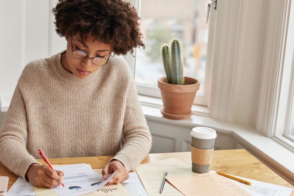 Concentrated statistician studies statistics, reads information data, examines chart, drinks coffee from disposable cup. Busy dark skinned architect works from home. People and documentation concept