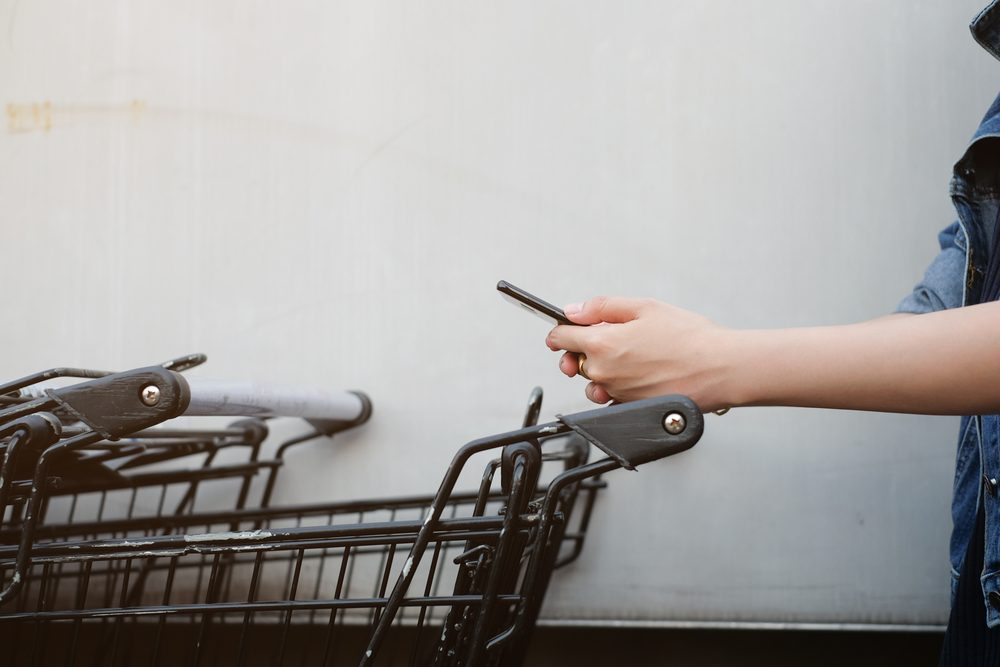 close up asian woman hand using smart phone and holding trolley bar for shopping on cement background at outside mall concept.