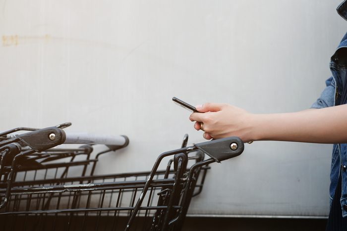 close up asian woman hand using smart phone and holding trolley bar for shopping on cement background at outside mall concept.
