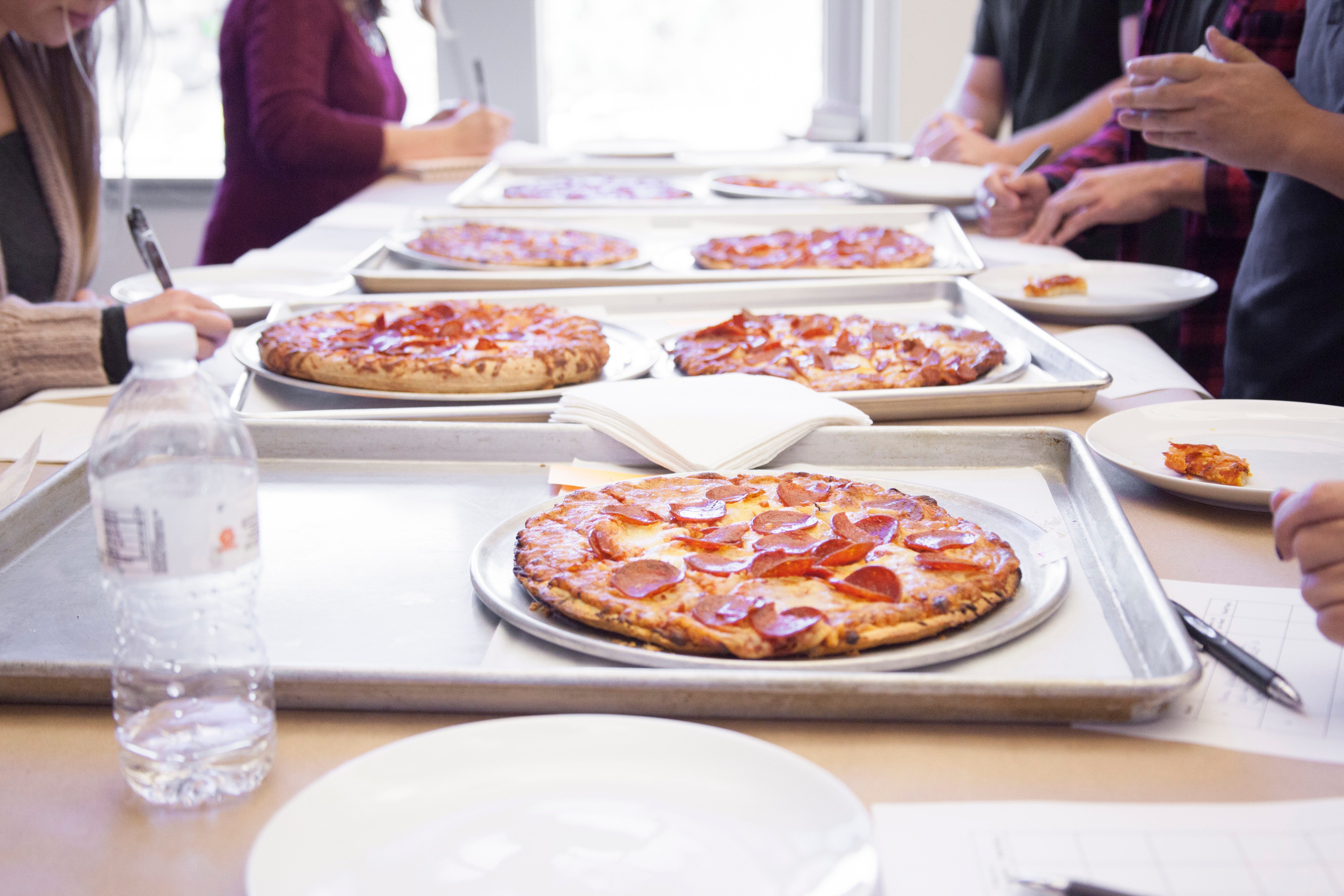 Table filled with unmarked pizzas with taste testers gathered around