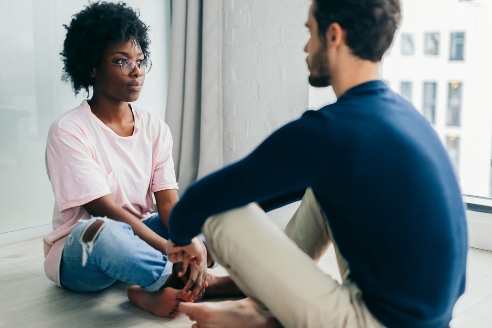 Young African woman and Caucasian man sitting near the window, staring at each other meditating together, free their minds from thoughts and worries.