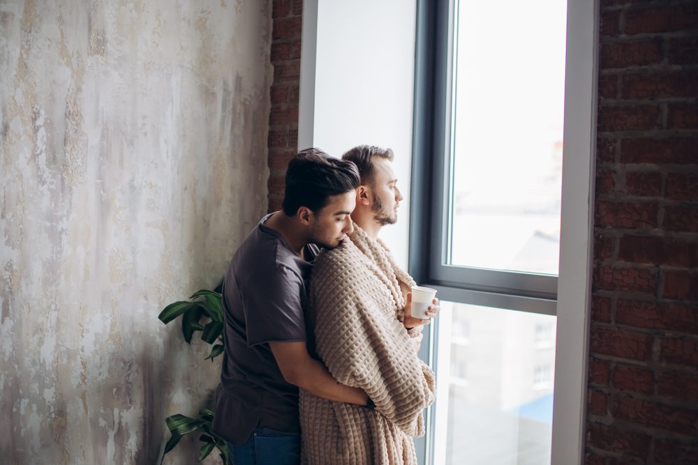 Romantic indoor shot of caucasian gay couple standing against window in modern loft design studio, looking at the street, dominant man taking care of his mate, tucking him with plaid