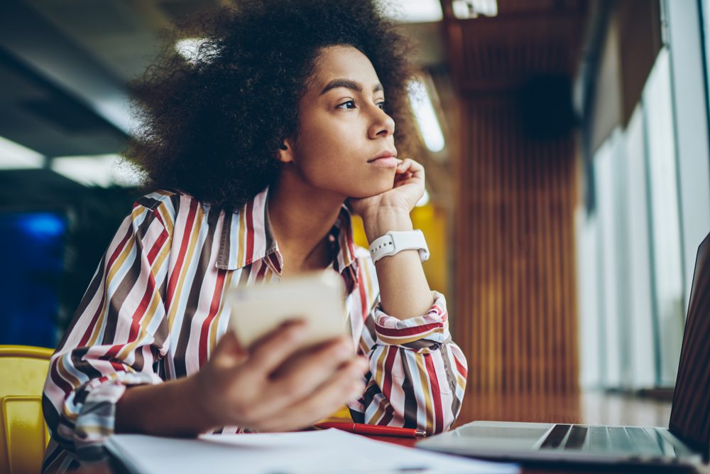 Pondering african american young woman thinking on creative ideas for publication in blog on smartphone device sitting in coffee shop.Thoughtful dark skinned hipster girl looking out of window