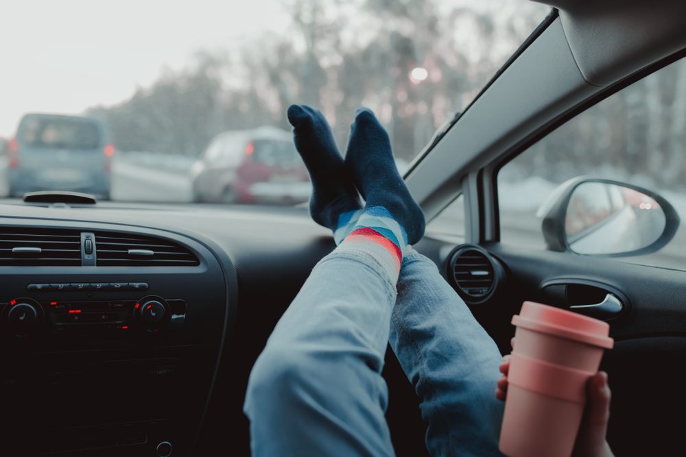 Woman is holding cup of coffee inside of car. Travel lifestyle. Legs on dashboard