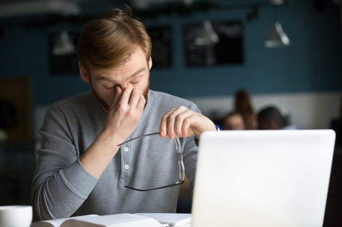 Young businessman taking off glasses feels eye strain tension tired of computer sitting at cafe table with laptop, millennial guy has bad sight vision problem massaging dry eyes after long laptop use