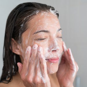 Skincare woman washing face in shower foaming facewash soap scrub on skin. Asian female adult cleaning body showering in hot water at home on in hotel as morning routine. Enjoying relaxing time.