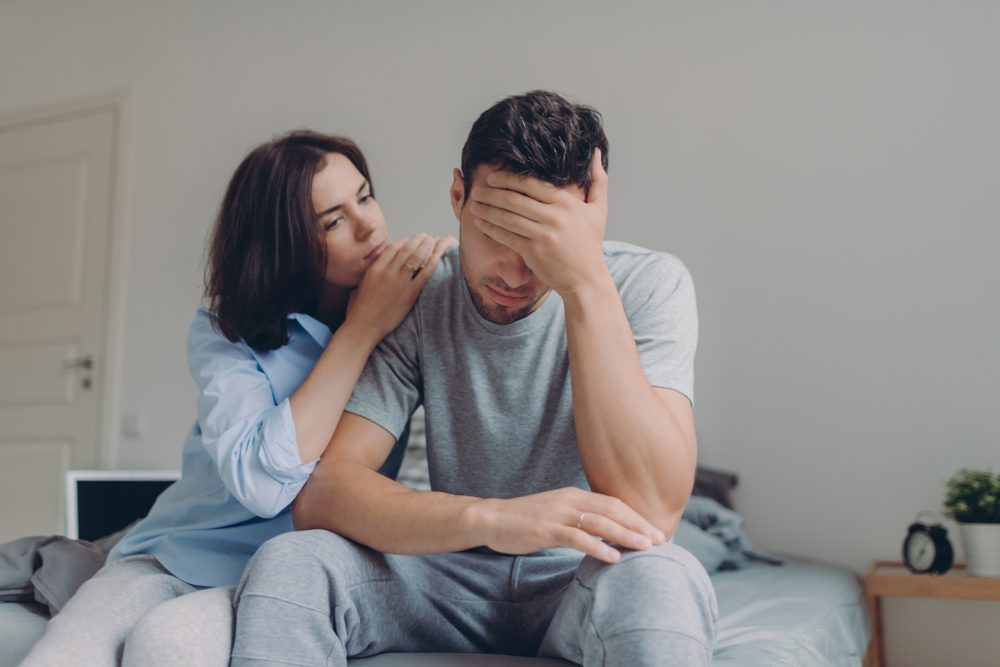 Attractive female consoles her sad boyfriend who has depression and some problems, pose at bedroom on bed, sit against domestic interior. Wife and husband try solve difficult situation in life.