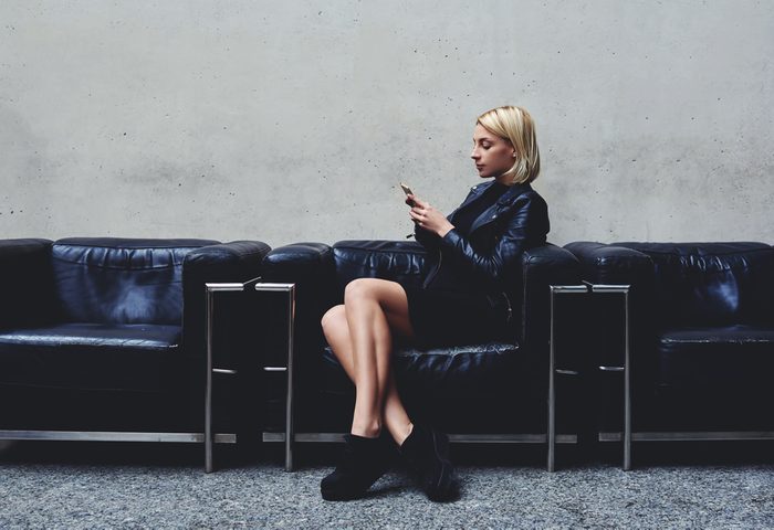 Stylish hipster girl chatting on mobile smart phone with her friend which she awaits before a photo session in the fashion studio, young blonde woman reviewing traffic in Internet via cell telephone