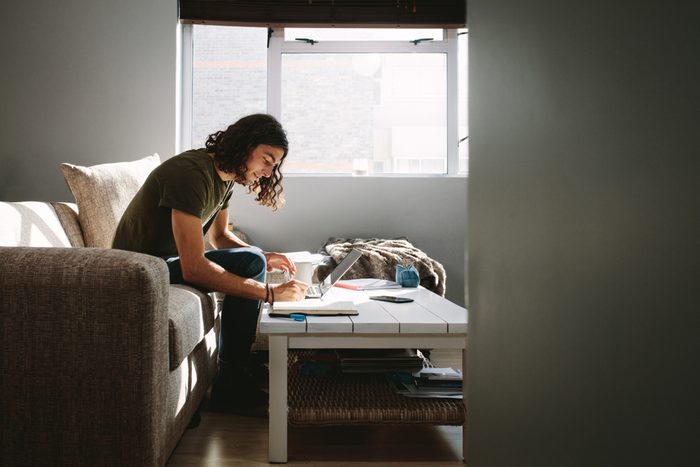 Student writing notes sitting on a couch beside a window at home. Young man studying with laptop and books on table.