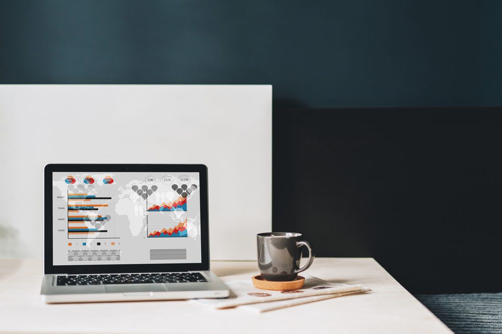 Workplace without people, close-up of laptop with graphs, charts, diagrams on screen on white table, desk.Nearby is closed paper notebook, pen, newspaper, cup of coffee. Online marketing, e-commerce.