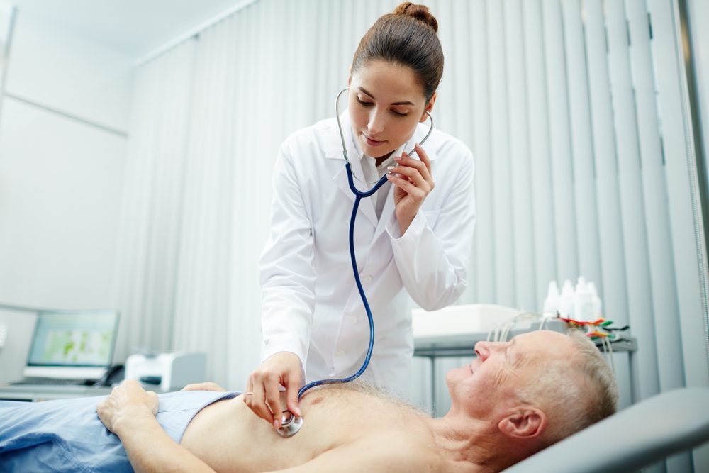 Serious concentrated attractive female doctor in lab coat using stethoscope to listen to sounds in lungs of elderly patient in procedure room